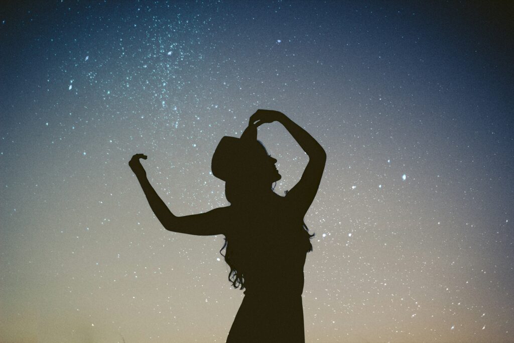 Silhouette image of a woman wearing a hat looking up towards the stars in a magical night sky.