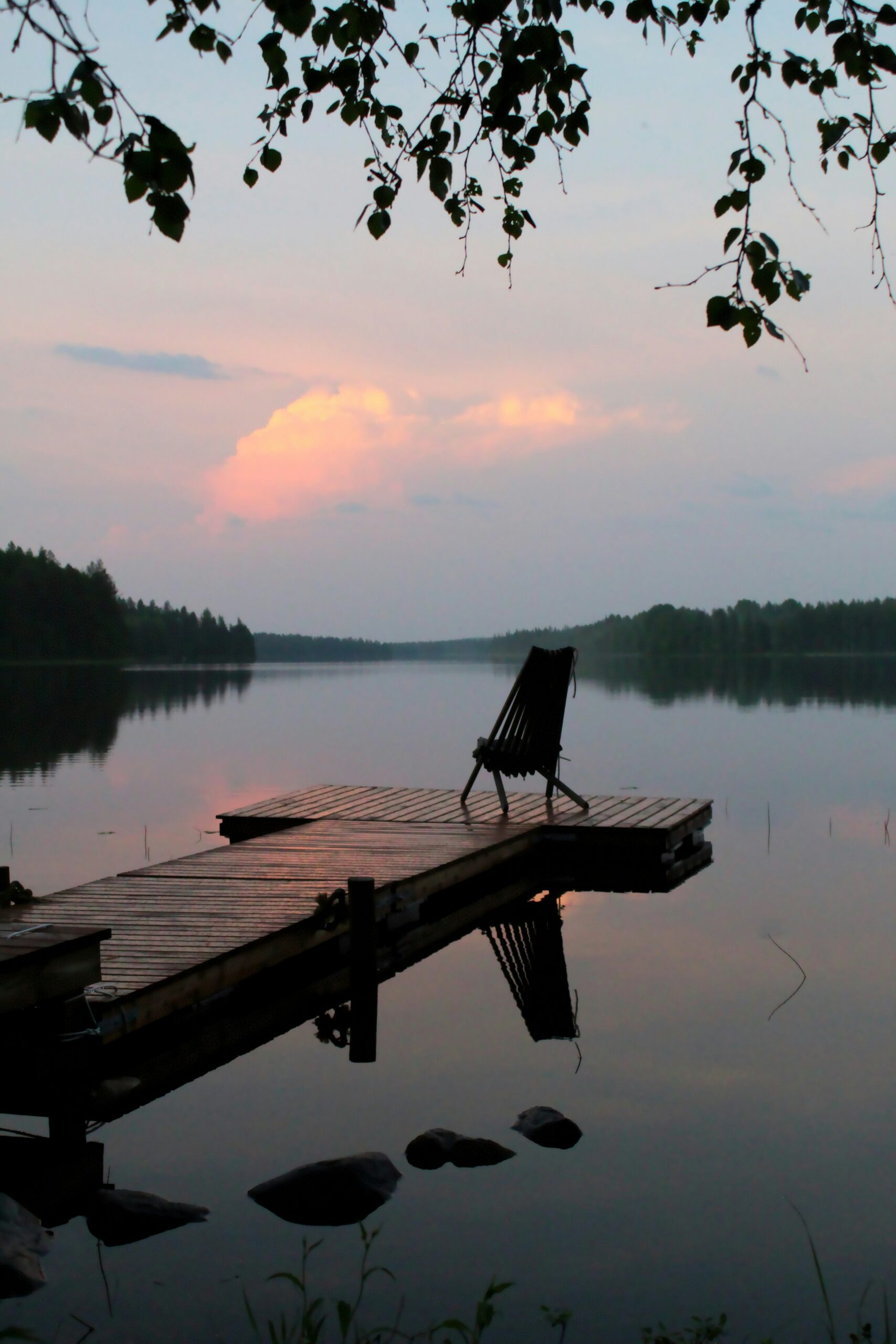 Picture of a chair on a jetty looking out on to a lake with a pink-orange sunset cloud wi