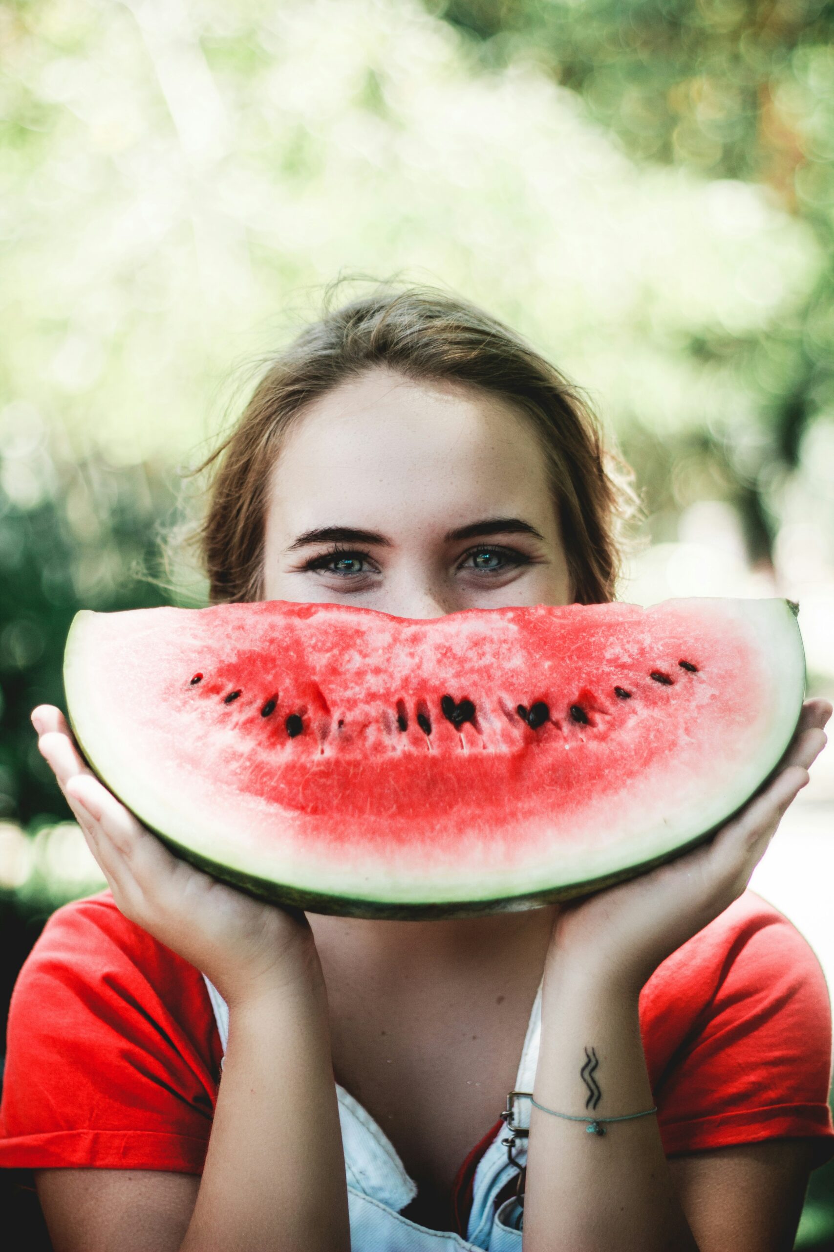 A picture of a woman wearing a red t shirt holding a melon up to her face in nature.