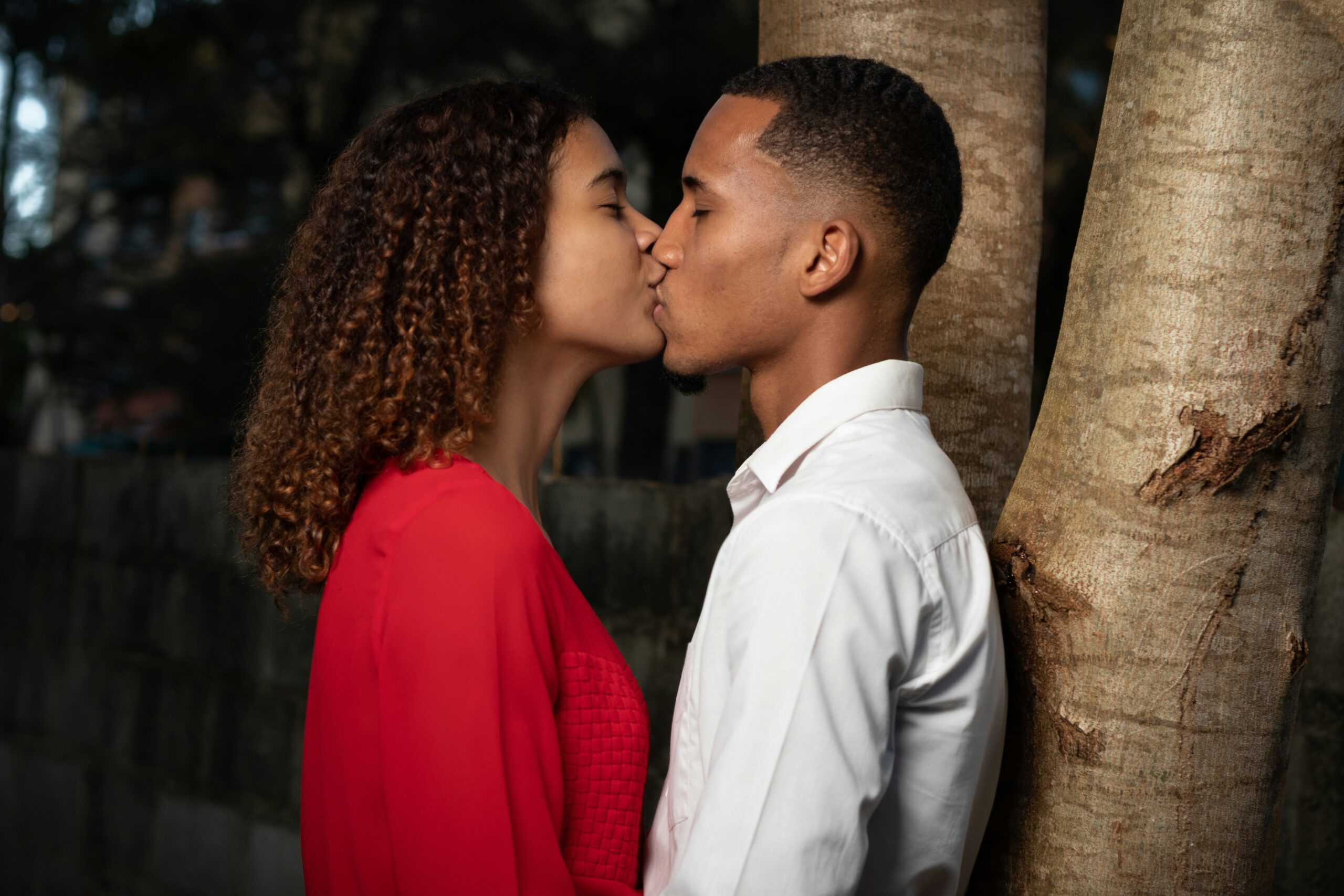 Photo of a lady wearing a red top and a man wearing a white top kissing beside a tree