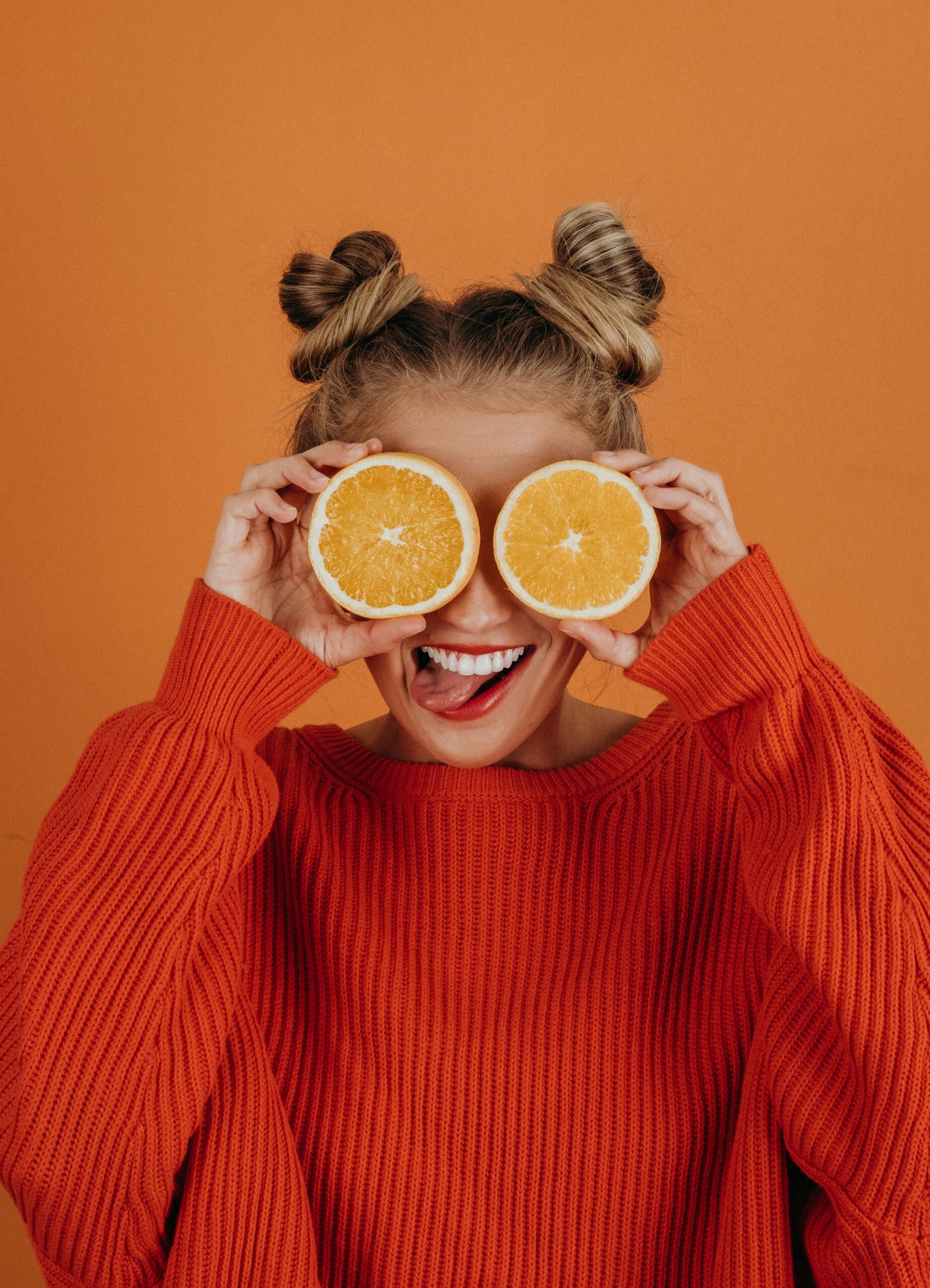 Women with hair in bunches wearing an orange jumper holding two oranges in front of her eyes to represent eyes on orange background.