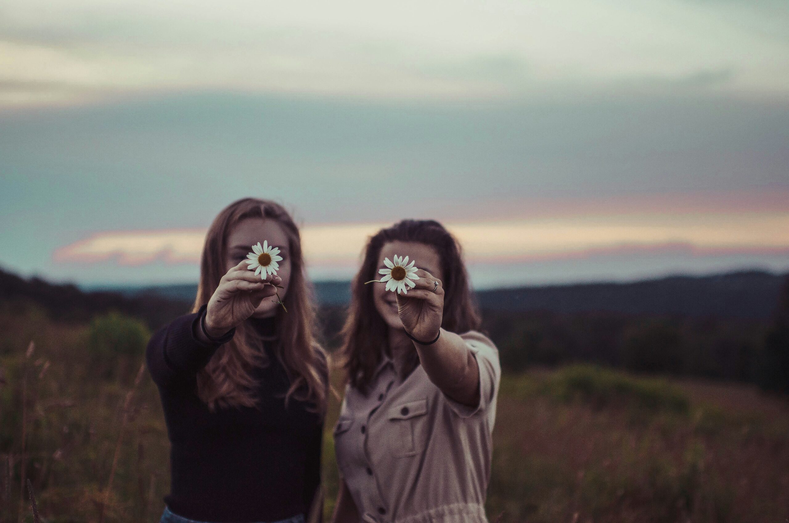 A picture of two young women with long hair in nature, both holding up large daisies.