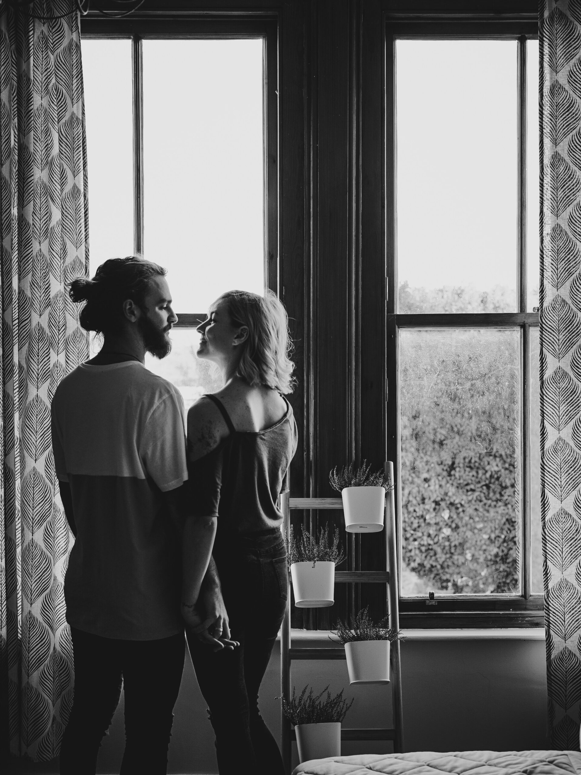 A black and white image of a couple in a house by the window looking into eachother's eyes