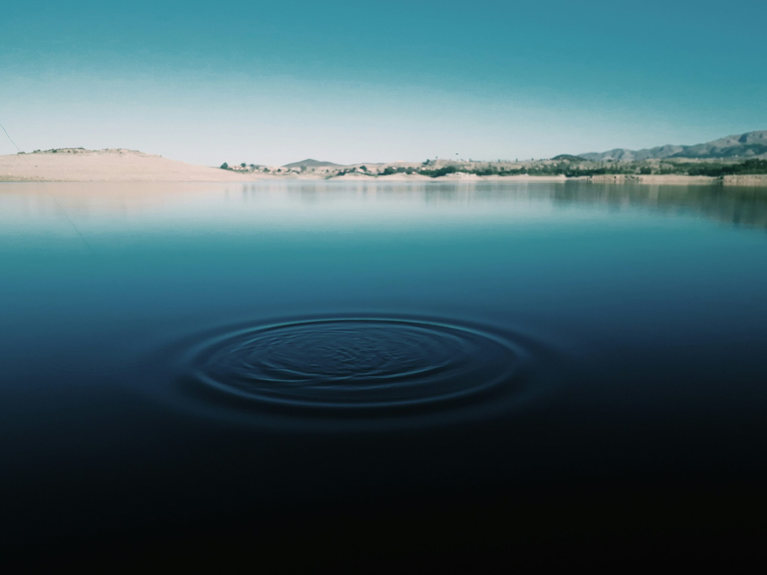 Alt Text: A serene lake stretches toward distant forested mountains under a bright blue sky. Gentle ripples ripple across the water’s surface, reflecting sunlight. The shoreline of smooth stones is visible in the foreground, and lush green trees frame the scene, evoking calm and natural beauty.